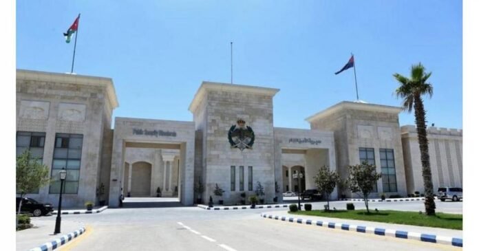 Front view of a beige stone government-style building with a central crest, three entrance arches, and flagpoles against a clear blue sky; a palm tree on the right and a curved driveway in the foreground.