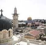 Aerial view of a historic Middle Eastern city with white stone buildings, domed roofs and minarets seen from above.