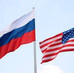 Russia and United States flags waving on flagpoles against a pale blue sky.