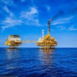 Offshore oil rig platforms standing in the deep blue ocean under a bright sky with a crane visible on one platform.
