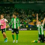 Soccer players in green-and-white uniforms celebrate on a bright stadium field, with one kneeling and pointing to the sky as teammates smile nearby.