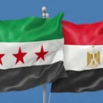 Two national flags waving against a blue sky: Egypt on the right and a green‑white‑black flag with three red stars on the left, both on flagpoles.