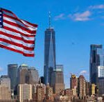 American flag waving above the Manhattan skyline with tall buildings against a blue sky, sunny day.]?