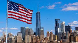 American flag waving above the Manhattan skyline with tall buildings against a blue sky, sunny day.]?