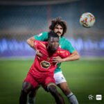 Two soccer players battle for the ball in a tight challenge on the field, one in red and one in green.