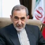 Older man with gray hair and beard in a dark suit seated in an office, with an Iranian flag in the background.