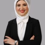 Professional portrait of a smiling woman wearing a white hijab, white blouse, and black blazer with arms crossed against a gray background.