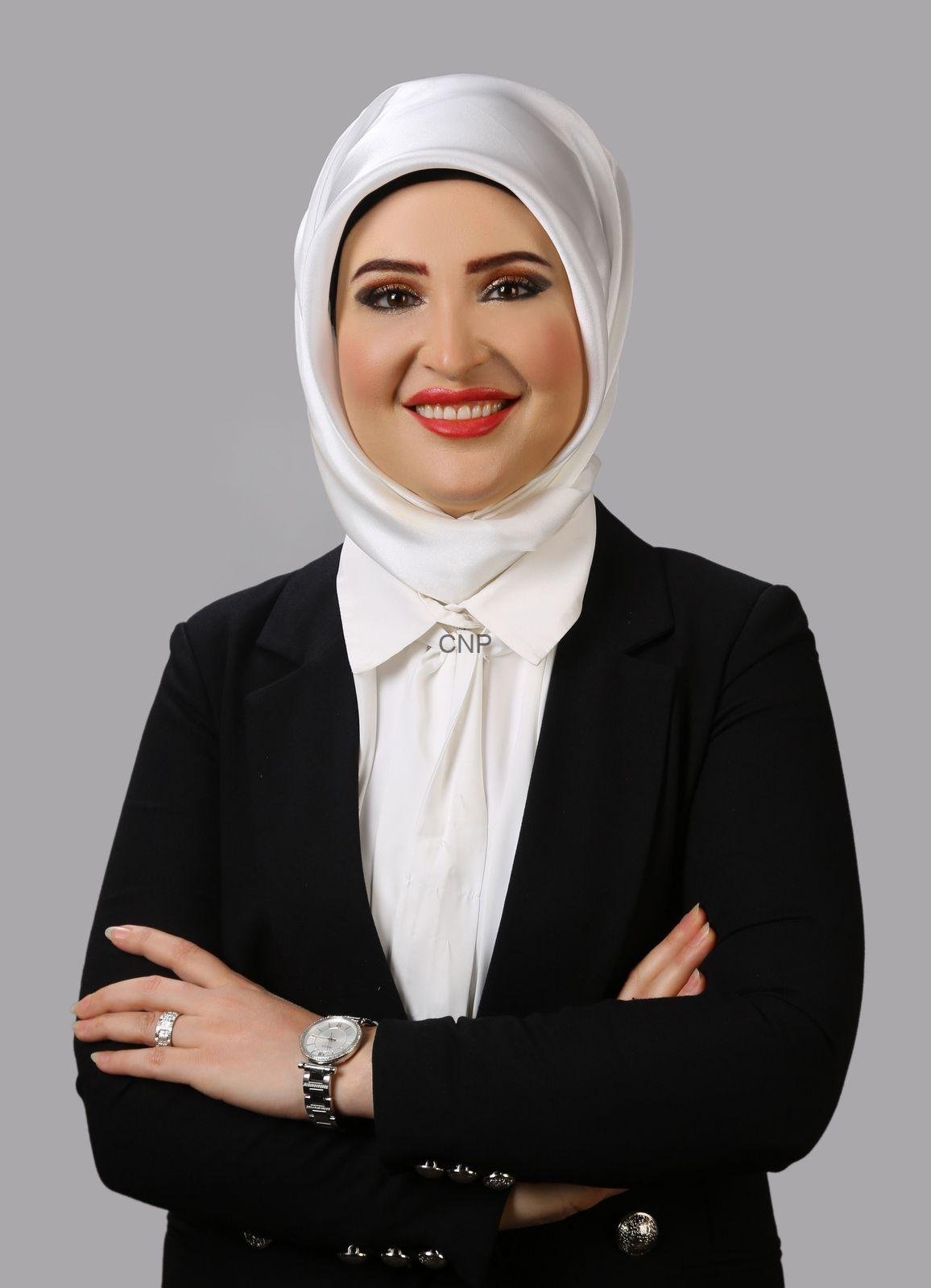 yas - Center News Network Professional portrait of a smiling woman wearing a white hijab, white blouse, and black blazer with arms crossed against a gray background.