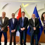 Five businesspeople in suits stand in a diplomatic office with Jordan, Turkey, and EU flags behind them.