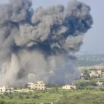 Large gray smoke plume rising from a rural village as buildings sit in open fields nearby (explosion in progress).