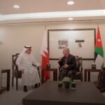 Diplomatic meeting in a formal room with four men seated, flags behind them and a central coffee table with plants.