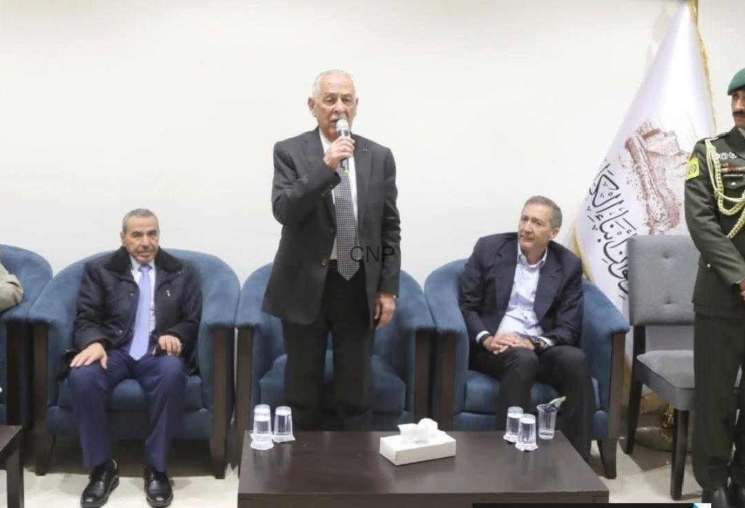 Man in a dark suit speaks into a microphone at a formal meeting, flanked by seated officials.