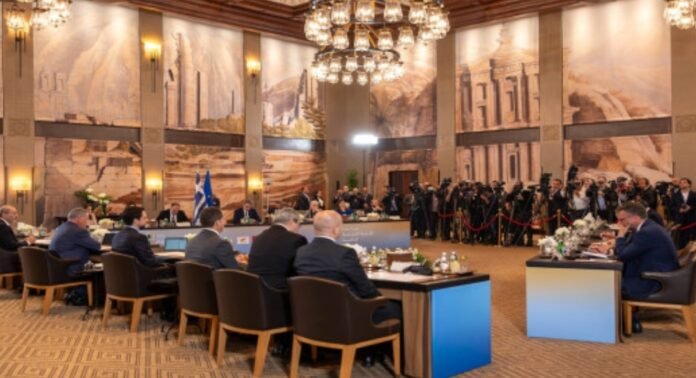 Delegates seated around a U-shaped conference table in a grand hall with murals, chandeliers, and press photographers at the back.