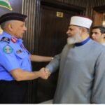 Police officer in a blue uniform shakes hands with a bearded man in a gray traditional robe and white cap as others stand in line nearby.