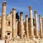 Row of tall, weathered stone columns from an ancient ruin, with stone walls and steps beneath a blue sky.