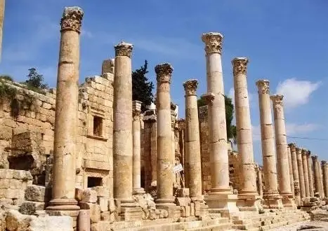 Row of tall, weathered stone columns from an ancient ruin, with stone walls and steps beneath a blue sky.