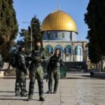 Group of soldiers in tactical gear standing in a stone courtyard with the Golden Dome of the Rock in Jerusalem in the background.