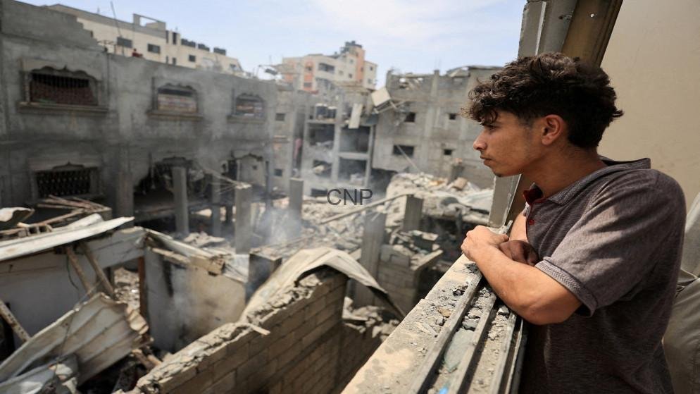 6a097cef2026d - Center News Network Young man leaning on a railing, gazing over a ruined urban landscape with collapsed buildings and smoke in the distance.