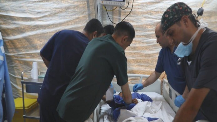 Medical team performing an emergency procedure at a hospital bed inside a tent-like field setup, with gloves and masks on.