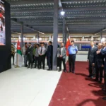 Group of officials and workers listening to a presentation inside a factory, with Jordan and China flags visible on stands to the left and center.