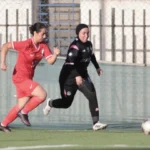 Two female soccer players chase a ball on an outdoor field; one in a red kit, the other in black with a hijab, near the fence and goal area.