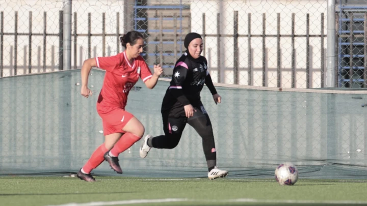 Two female soccer players chase a ball on an outdoor field; one in a red kit, the other in black with a hijab, near the fence and goal area.
