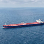 A large cargo ship sailing across a calm blue ocean, viewed from above.