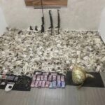 Rifles propped against the wall with bundled cash, a scale, and ammunition on the floor, implying illicit activity.