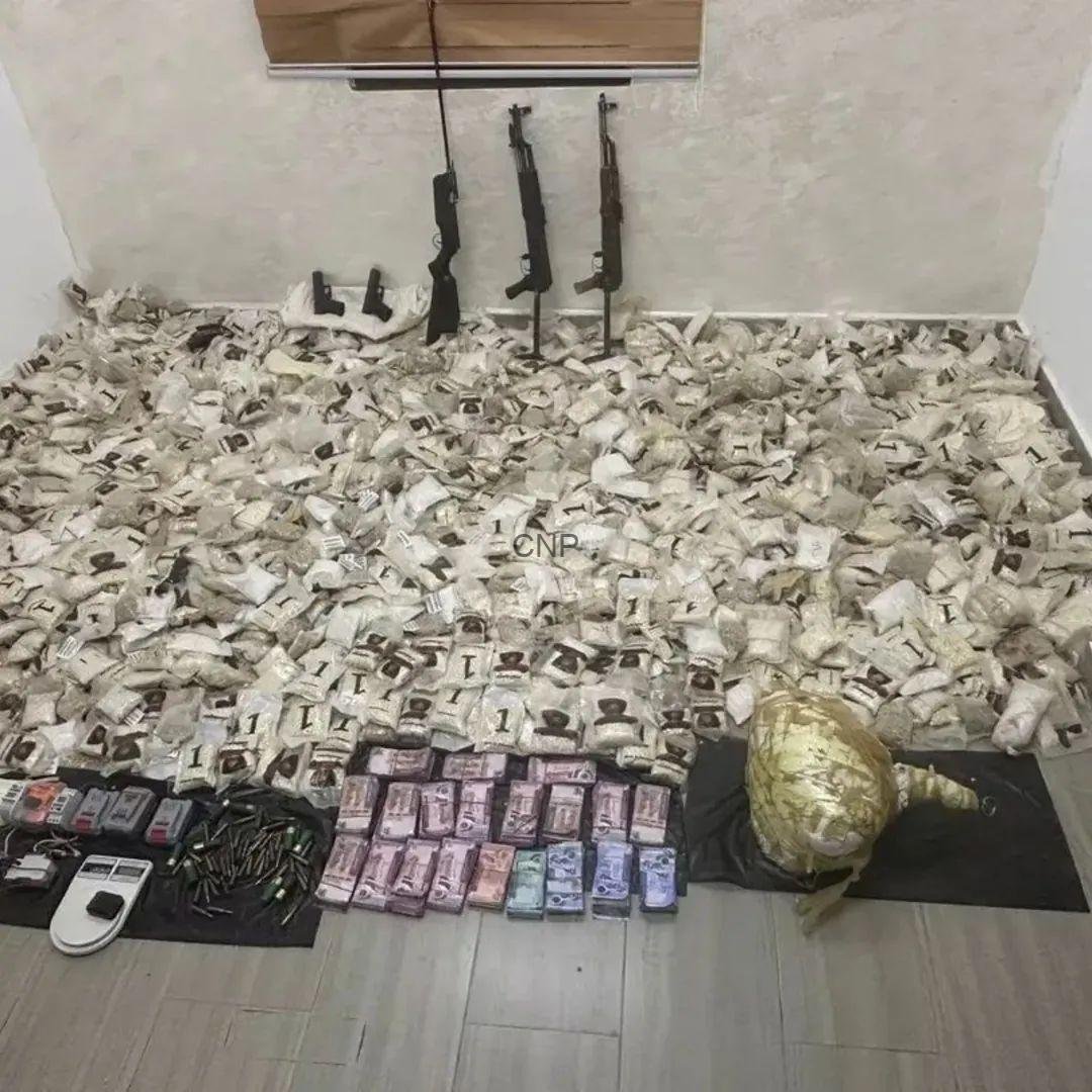 Rifles propped against the wall with bundled cash, a scale, and ammunition on the floor, implying illicit activity.