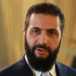 Man with a dark beard and navy suit looking at the camera in a softly lit room.