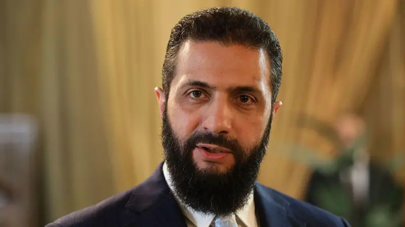 Man with a dark beard and navy suit looking at the camera in a softly lit room.