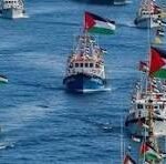 Several small boats display tall colorful flags as they float on a bright blue sea in a harbor or open water area.