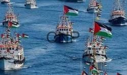 Several small boats display tall colorful flags as they float on a bright blue sea in a harbor or open water area.