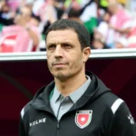 Athletic coach standing on a sideline, wearing a black Kelme jacket with a Jordan crest, with a blurred stadium crowd in the background.