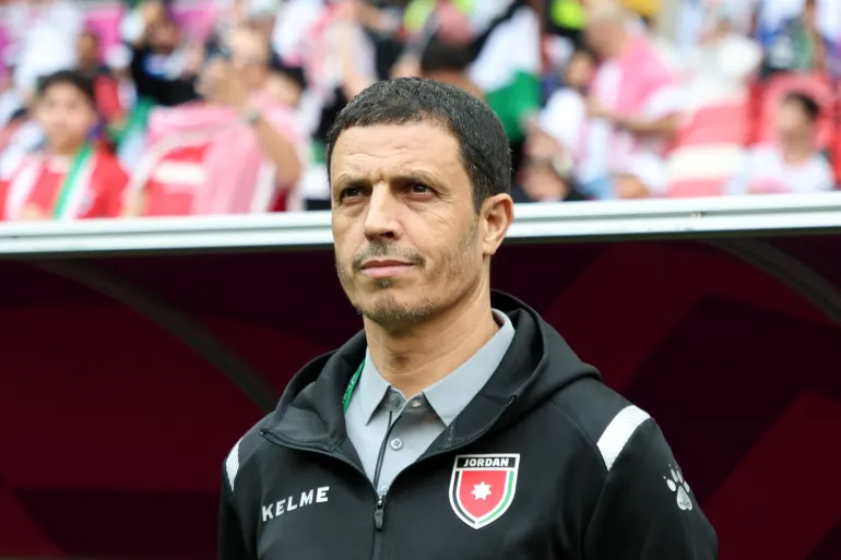- Center News Network Athletic coach standing on a sideline, wearing a black Kelme jacket with a Jordan crest, with a blurred stadium crowd in the background.