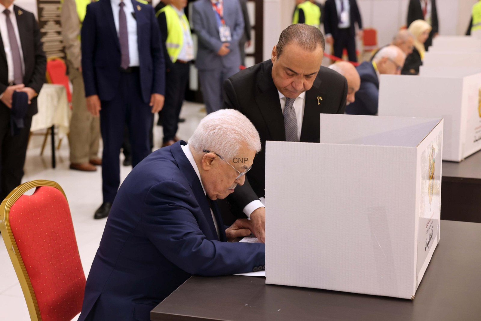 Man in a dark suit signing a ballot at a polling booth, with another man overseeing nearby