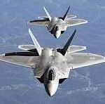 Two gray military fighter jets fly in close formation over a mountainous landscape, viewed from above.