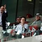 Group of people standing on a glass-front balcony, some raising hands and cheering in a crowded venue.