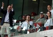 Group of people standing on a glass-front balcony, some raising hands and cheering in a crowded venue.