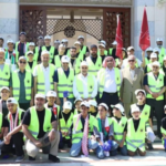 Group photo of volunteers wearing neon safety vests, kneeling and standing in front of a building with red flags in the background.