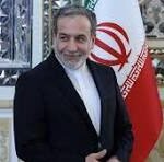 Man in a dark suit standing indoors beside the Iranian flag, smiling, with ornate wall decor in the background.