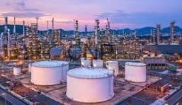 Oil refinery at dusk with large white storage tanks, pipes and processing towers, and a distant city skyline.