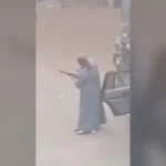 A person in a long robe aiming a rifle beside an open car door in a street scene.