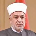 Religious figure wearing a white turban and glasses, speaking at a formal event with red flags in the background.