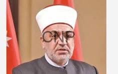 aw - Center News Network Religious figure wearing a white turban and glasses, speaking at a formal event with red flags in the background.