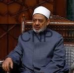Man wearing a white kufi cap and dark traditional outfit seated in a patterned chair in a formal setting.