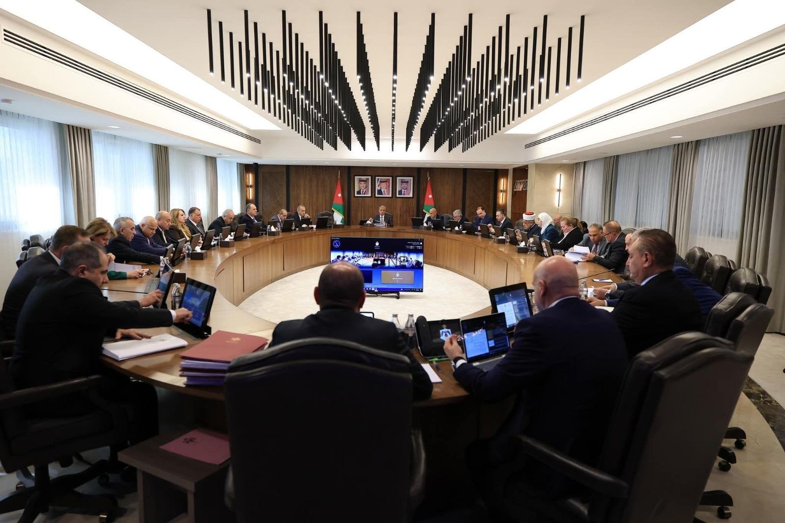 Formal government meeting: officials seated around a large curved table with laptops, central screen, and Jordanian flags in the background.