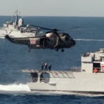 Military helicopter hovering above a gray naval ship at sea with a Union Jack on its side