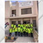 Group of people in yellow safety vests posing in front of a beige building with an Arabic banner above the entrance.