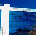 Modern glass-fronted office building with blue-tinted windows, two flagpoles bearing flags, and a clear blue sky above.